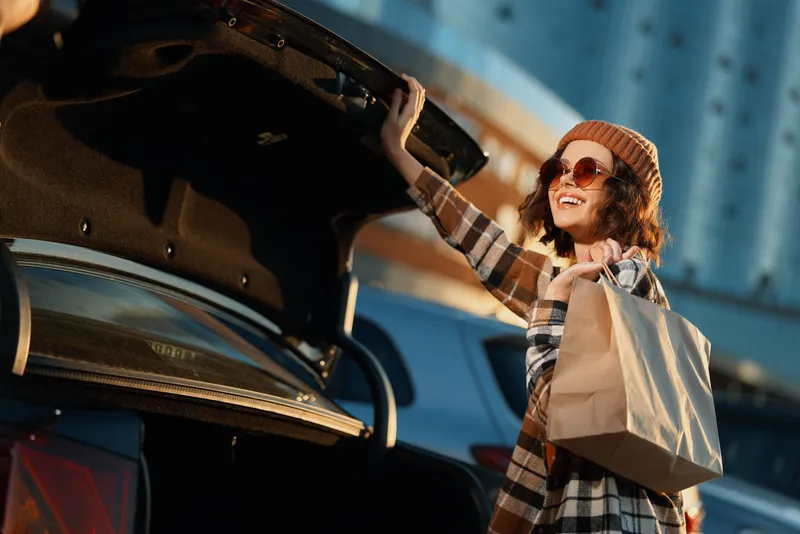 woman car trunk shopping smile urban scene with sunlight and golden glow, candid lifestyle portrait of a happy shopper in coat and beanie, authenticity, golden hour glow, mindful living and emotional