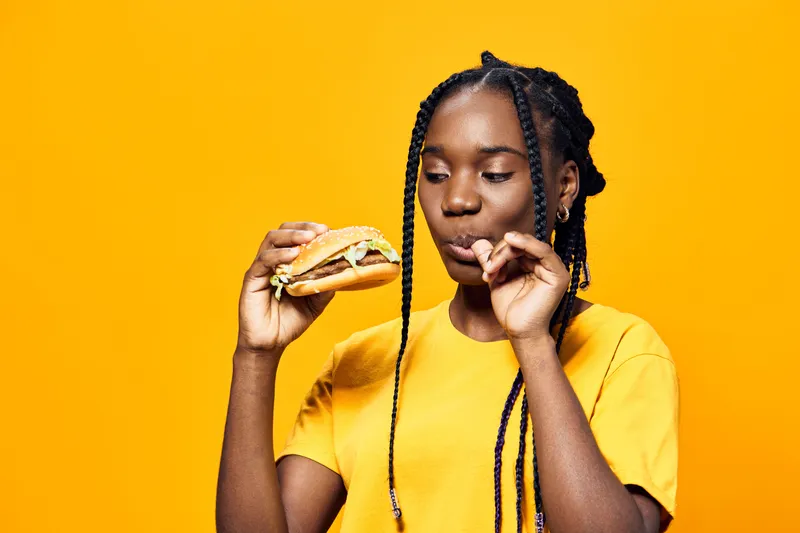 Young girl enjoying a delicious burger, wearing a bright yellow shirt, set against a vibrant yellow background, showcasing joy and a love for food