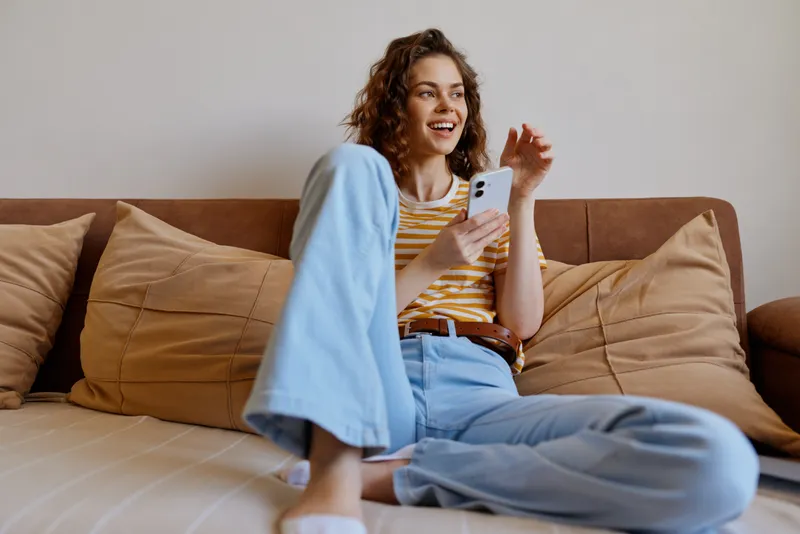 Relaxed young woman sits on a soft bed at home, wearing a striped top and light blue jeans, scrolling on her smartphone with a bright, genuine smile, surrounded by warm pillows and calm atmosphere