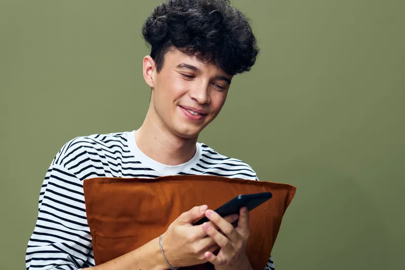 young man smiling while looking at smartphone, holding brown pillow, curly hair, casual striped shirt, indoor, olive green background