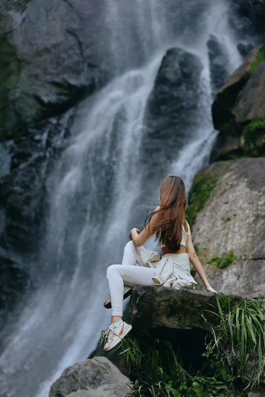 woman sitting near waterfall wearing white clothes with long hair in natural green rocky outdoor landscape, peaceful and relaxing atmosphere