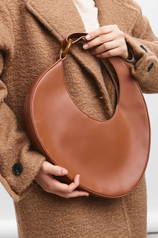 asian woman wearing a tan coat presents a circular leather handbag in a fashion forward studio shot. The image emphasizes texture and chic simplicity, creating a refined lifestyle impression.