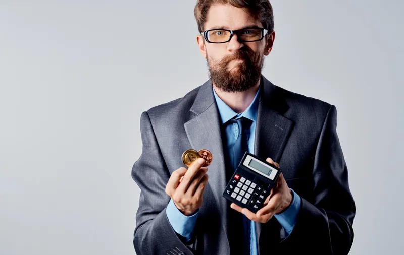 Мужчина в деловом костюме держит калькулятор и монету криптовалюты Bitcoin