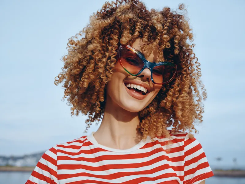 Bright portrait of a joyful woman with voluminous curly hair, wearing vivid sunglasses and a red striped tee, bathed in warm outdoor sunlight and a cheerful vibe.