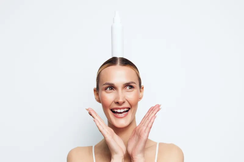 Smiling woman in studio portrait, hands near face, fresh skincare routine concept, serum bottle above head, beauty and spa mood, clean white background, soft light, calm expression
