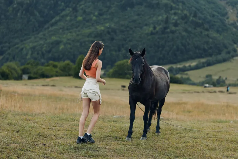 A young woman in a sleeveless top and shorts interacts with a horse in a sunlit meadow beneath rolling hills. The scene conveys calm, freedom, and nature.