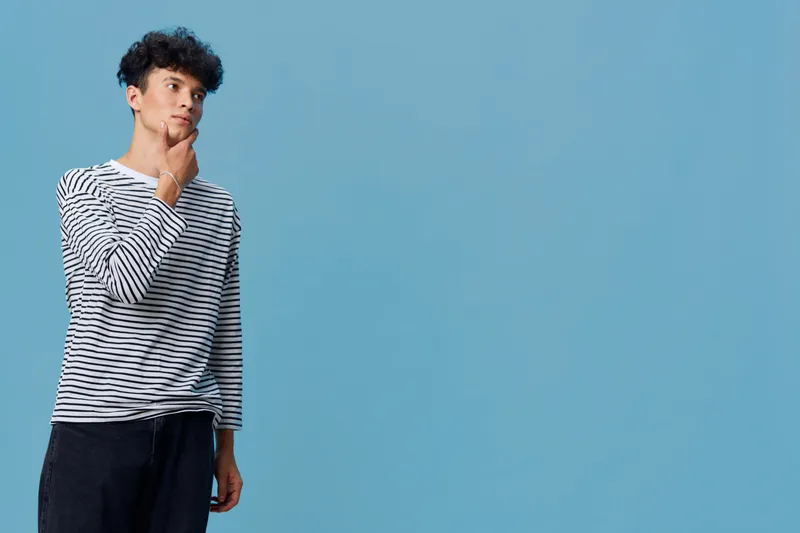 thoughtful man wearing striped long sleeve shirt stands against blue background, looking away with hand on chin, casual style, young adult, pensive mood