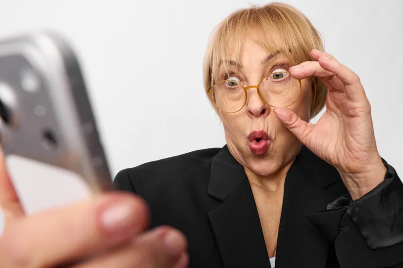 Elderly woman with glasses holding them near her face, surprised expression as she takes a selfie on a smartphone, portrait, soft white studio light, home technology