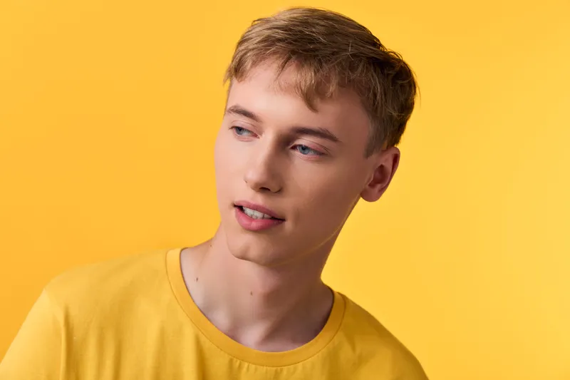 Young man in a yellow shirt stands against a bright yellow backdrop, looking to the side with a calm expression, casual fashion, studio lighting, clean composition, lifestyle image for advertising