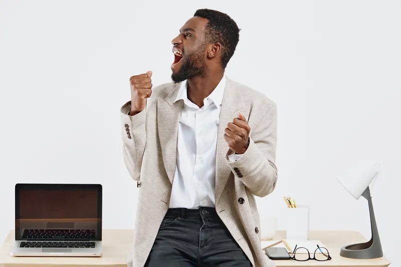 Enthusiastic smiling man in smart casual outfit celebrates success at his modern workspace with laptop and accessories on a white background, expressing happiness and motivation. Business lifestyle co