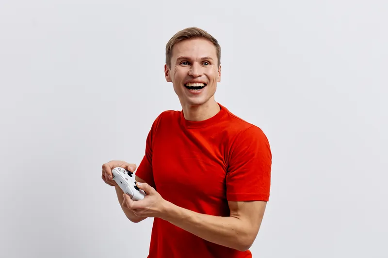 Smiling man in red t-shirt holding game controller, looking excited and enthusiastic, playing video game, indoor, casual wear, joyful expression, entertainment concept, studio shot.