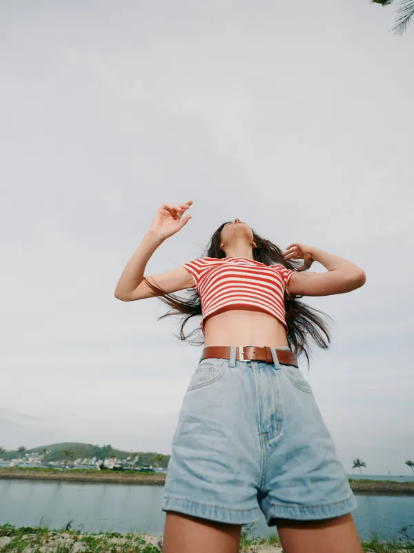 Low angle shot of a person in a striped cropped top and denim shorts posing outdoors by a calm body of water under a pale sky, capturing bold style and carefree attitude.