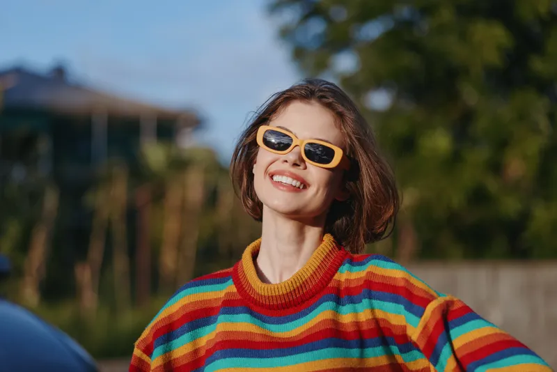 Woman smiling in a colorful striped sweater and yellow sunglasses, young adult enjoying sunlight outdoors. Casual outfit, short hair, joyful travel lifestyle portrait in green suburban setting.