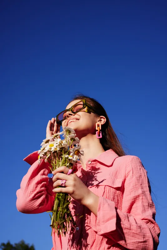 Beautiful young woman with long hair enjoying the sunshine, wearing a pink shirt and holding a bouquet of daisies, with a blue sky background
