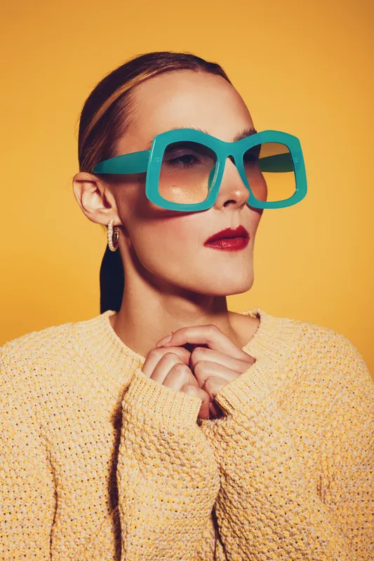 Stylish woman with oversized turquoise sunglasses and red lipstick poses confidently against yellow background wearing cozy knitted sweater. Concept of fashion, beauty, and bold accessories.