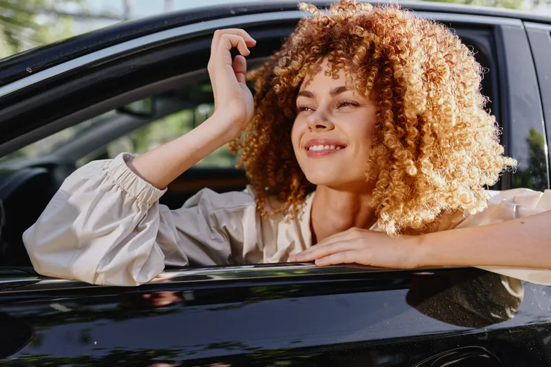 Smiling woman with curly hair leans out of car window, enjoying sunlight and fresh air, wearing light blouse on a bright sunny day in an outdoor setting.