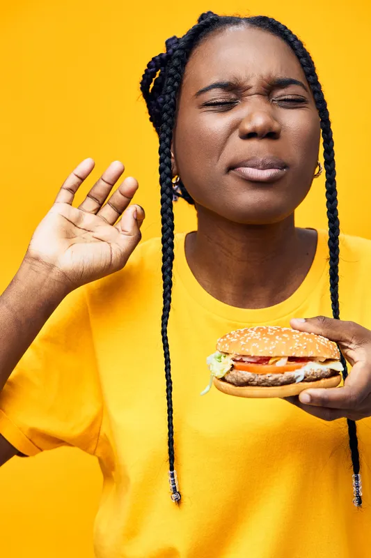 A young woman of African descent, enjoying a delicious burger with a joyful expression, set against a vibrant yellow background, perfect for food related concepts