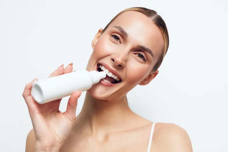 Smiling woman with clear skin holding white cosmetic bottle near mouth on white background, demonstrating skincare product application. Bright lighting highlights fresh and healthy appearance.