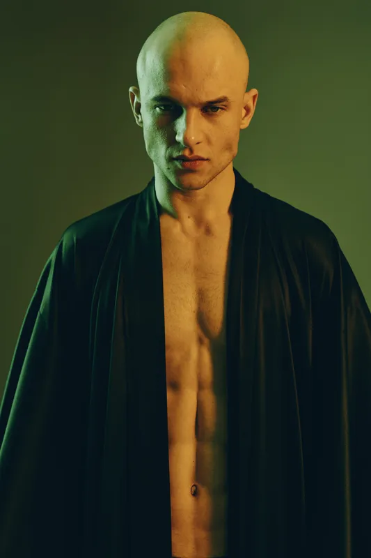 Handsome man poses confidently in a sleek black robe, showcasing a strong physique and intense expression against a warm, ambient background.