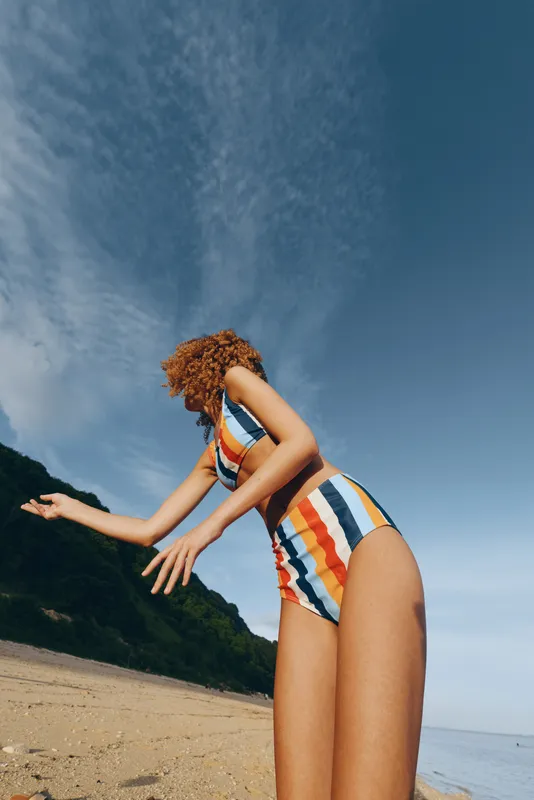 Young woman with curly hair enjoys a sunny day on the beach wearing a colorful striped swimsuit under a partly cloudy sky.