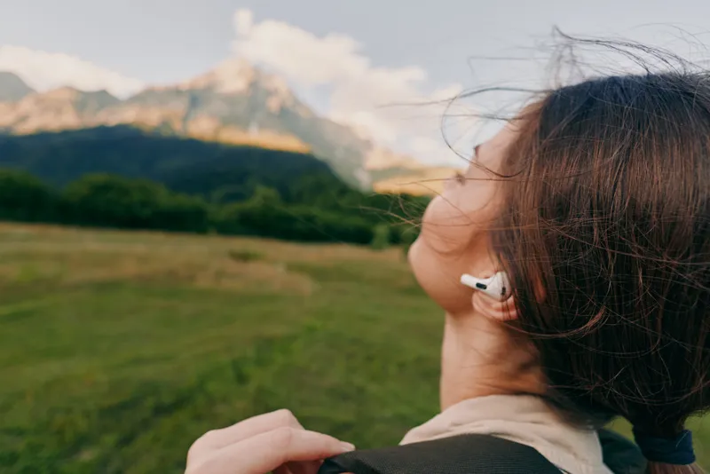 Woman outdoors with backpack, earphones, in a sunlit field near mountains, capturing a relaxed adventurous lifestyle moment in natural colors and fresh air
