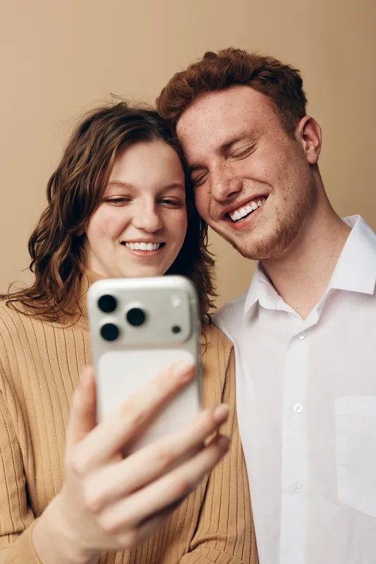 Smiling young couple takes selfie with smartphone, close together in casual clothes on beige background. Concept of friendship, technology, happiness, and modern lifestyle.