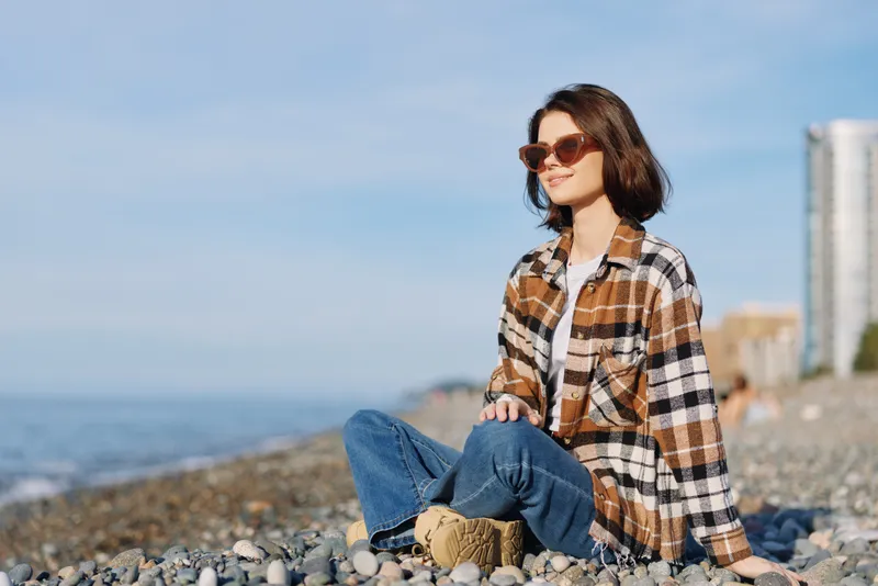 Woman on a pebble beach wearing a plaid shirt, jeans, and sunglasses, relaxing by the sea with a calm expression and breezy outdoor fashion vibes.