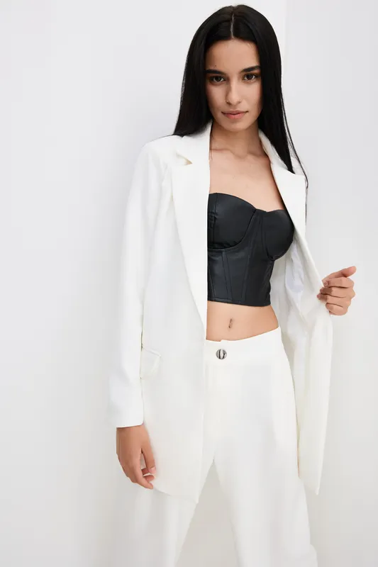 Woman wearing white suit jacket and pants with black leather corset top, standing confidently against white background in minimalistic studio setting, stylish modern outfit.