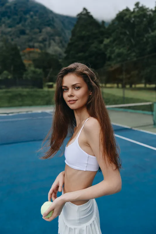 Young athletic woman in a sporty outfit with long hair, holding a tennis ball on a blue court surrounded by greenery