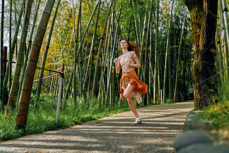 Woman running along a gravel path in a bamboo forest wearing a flowing skirt and blouse, joyful summer portrait of active lifestyle, motion, sunlight and serene outdoor nature.