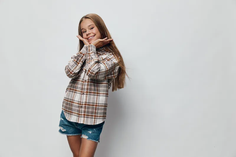 Girl smiling and posing with hands near face wearing plaid shirt and denim shorts in studio. Happy child with long hair standing with casual clothes on grey background.