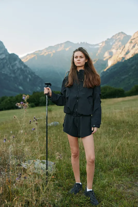 A poised outdoorswoman stands in a scenic alpine meadow with a trekking pole, wearing a dark jacket and shorts. She gazes ahead with confidence and focus.