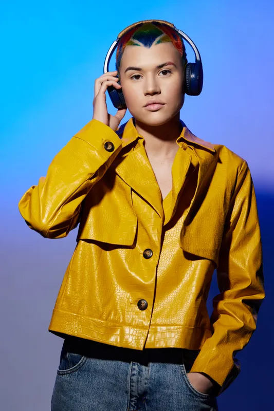 young woman with colorful hair wearing yellow jacket and headphones, standing against gradient blue-purple background, modern urban style, casual outfit.