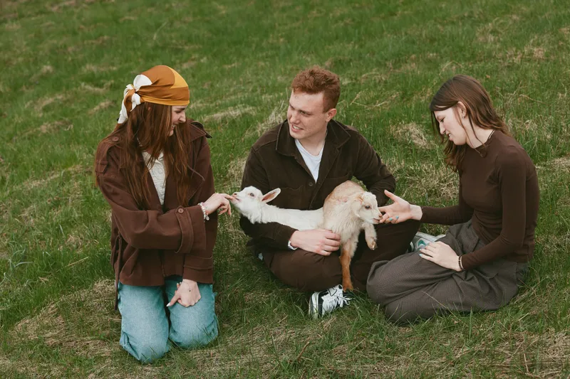 Three young adults sit on grass in casual clothes enjoying a sunny day while holding and petting a baby goat in a relaxed outdoor lifestyle setting.
