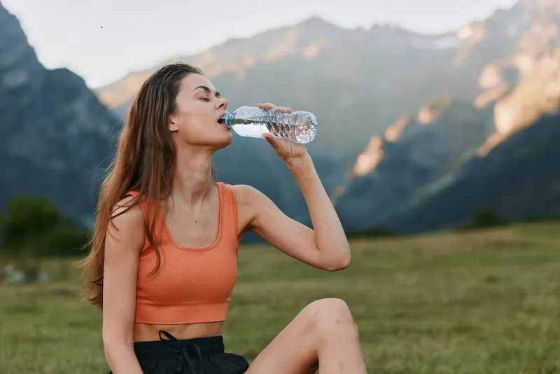 A woman in an orange sports top drinks water from a bottle in a scenic mountain meadow. She sits on the grass with distant peaks, enjoying hydration during outdoor activity.