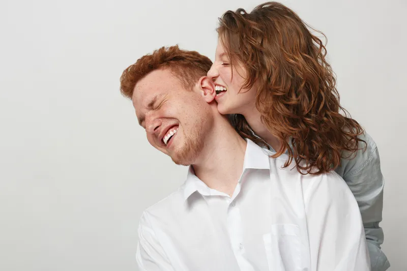 Happy young couple with curly hair laughing and enjoying playful moment together. Man and woman in white shirts share joyful intimacy on plain light background. Concept of love and fun.