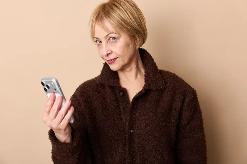Senior woman in studio portrait, surprised expression while holding smartphone, beige background, indoors communication and technology theme, hands touch screen, calm mature style