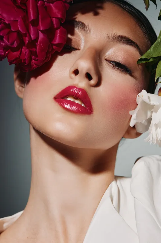 Creative fashion beauty portrait of woman with glossy red lips and floral accessories against a soft gradient background. Studio shot emphasizing makeup and style details.