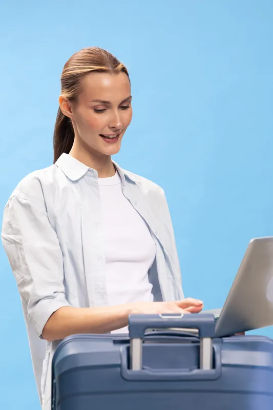 Young woman with suitcase using laptop on blue background. Travel concept with casual clothing and positive expression preparing for trip or vacation planning.
