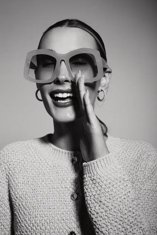 Happy caucasian woman with fair skin and large glasses laughs joyfully, wearing a knitted sweater against plain background. Concept of fashion, happiness, and lifestyle.