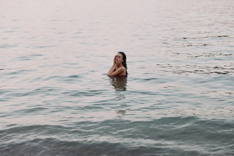 Smiling woman enjoying calm water at sunset, relaxing in gentle waves with peaceful expression and soft natural colors in background.