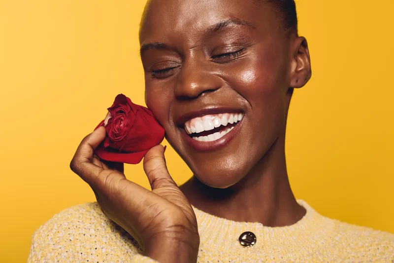Happy African woman with dark skin smiling with closed eyes holds red rose near face. Yellow background and beige sweater create warm mood. Concept of joy, beauty, and floral emotion.