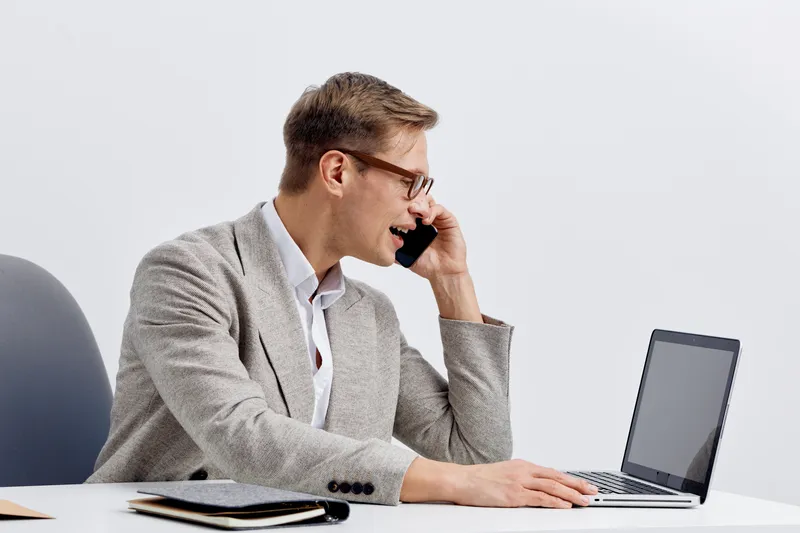 Businessman talking on mobile phone while working at his desk in modern office, using laptop, with documents and notepad, professional work environment, corporate lifestyle concept, full body, smiling