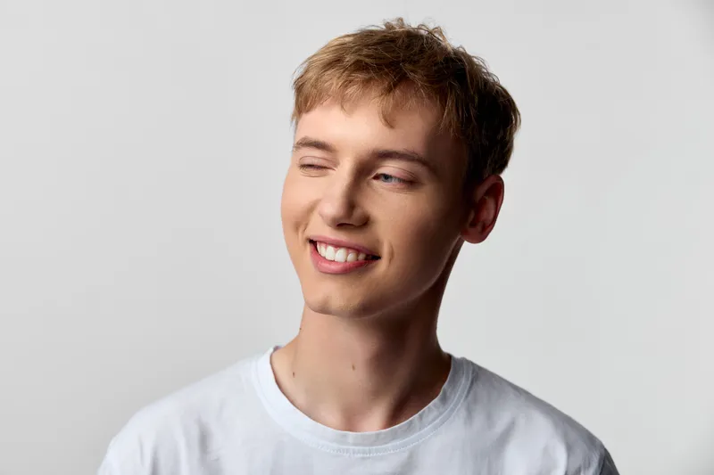 Smiling young man in a casual white tee, eyes closed with a warm expression, relaxed mood, soft studio lighting highlighting friendly vibe and everyday confidence and natural charm