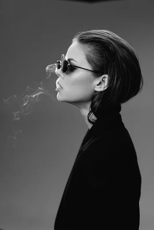 Black and white portrait of a stylish woman wearing sunglasses exhaling smoke in a dark minimalist setting.