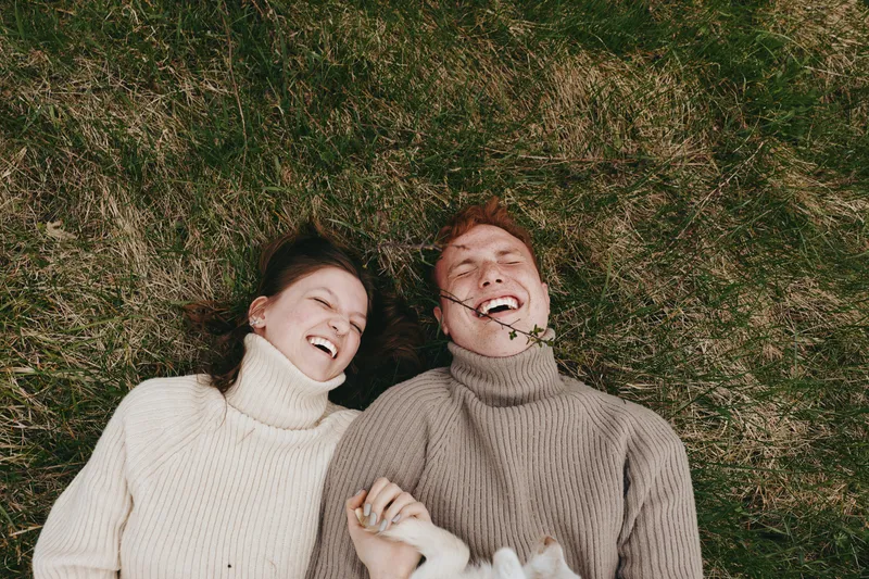 Two people lie on grass wearing turtleneck sweaters, sharing laughter and a relaxed moment outdoors, illustrating cozy lifestyle and genuine happiness