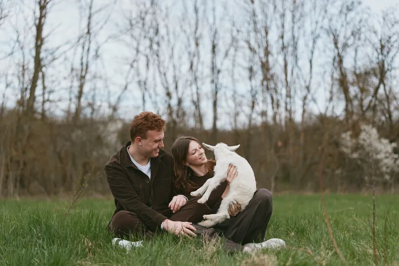Friendly couple relaxing outdoors with a playful goat kid, sharing smiles and gentle affection. Close-up on lush grass and bare trees, capturing lifestyle moments, wellness, and a warm, natural