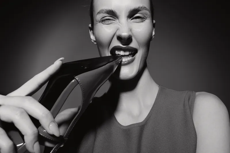 Young white woman with light skin biting a high heel shoe, playful expression on face, studio shot in black and white, fashion and fun concept.