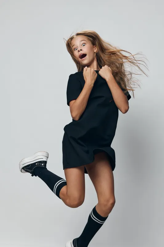 Girl jumping and smiling with excitement in casual black dress and sneakers, hair blowing and expression of joy on light background, youthful energy and motion captured.