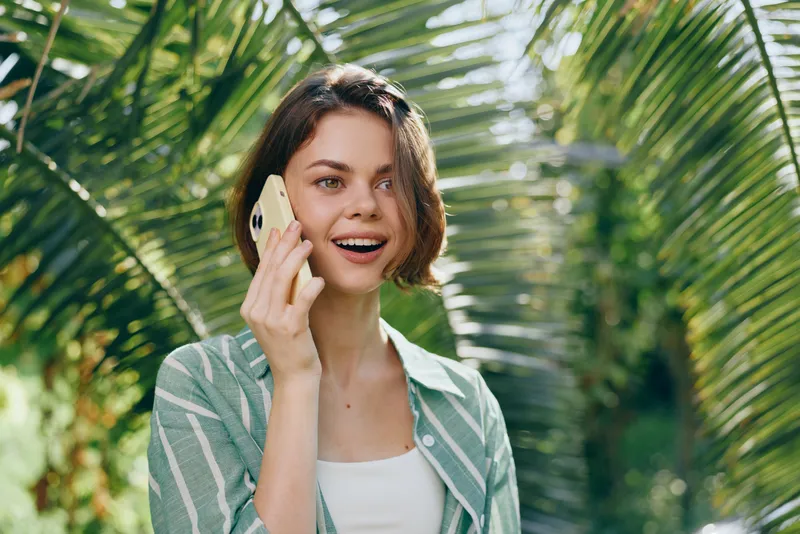 Smiling woman talks on a smartphone outdoors among lush palm leaves, bright daylight, casual attire, capturing a cheerful, everyday moment.